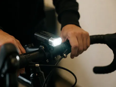 Vue rapprochée d'une lumière LED brillante fixée sur le guidon d'un vélo avec des mains ajustant le câble, sur un fond sombre et une paroi chaude.