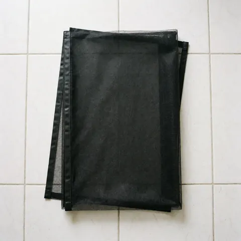Moustiquaire à fermeture magnétique noire, pliée sur un sol carrelé blanc, prête pour l'installation dans un intérieur.