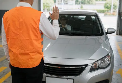 Un employé en gilet orange guide une voiture grise avec des gestes. Des flèches jaunes marquent le sol en béton près d'une entrée de garage.