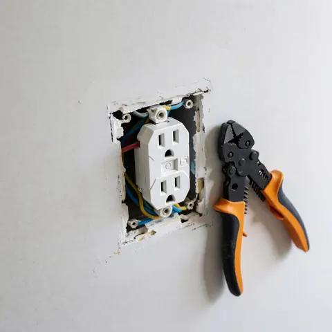 Une prise électrique blanche fraîchement installée sur un mur blanc, avec des câbles multicolores visibles et une pince à côté.