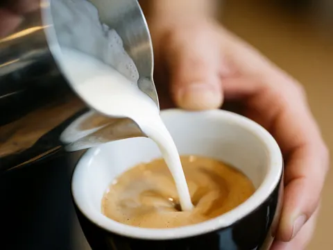 Lait versé depuis une carafe métallique dans une tasse noire d'expresso, créant un dégradé doux avec texture réaliste sur fond flou.