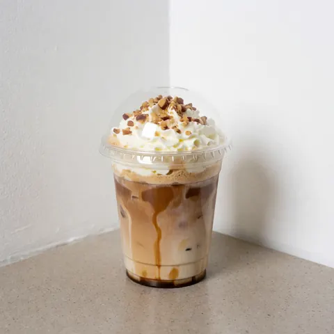 Verre transparent contenant du café glacé, garni de crème fouettée et de noix hachées, posé sur une surface en pierre claire contre un fond blanc lumineux.