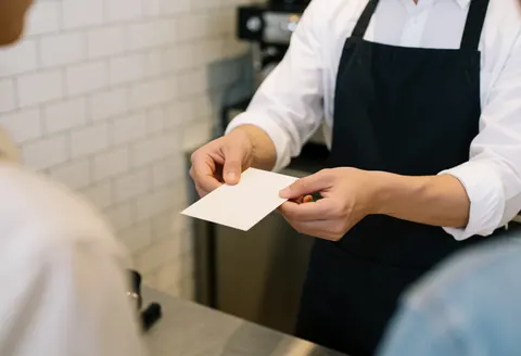 Un barista montre un menu blanc à un client dans un café. Ses mains et les outils sont visibles sur un fond de cuisine professionnelle avec des carreaux blancs.