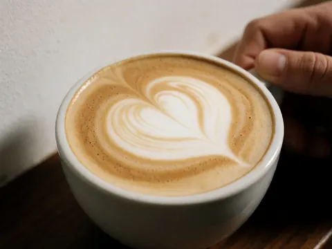 Tasse blanche contenant un cappuccino avec un motif en lait en forme de cœur, posée sur une table en bois sombre. Des mains tiennent la tasse sur fond blanc.