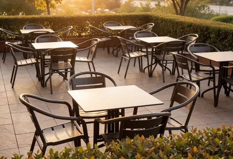 Terrasse de café au coucher du soleil avec tables et chaises en métal, sol en pierre et haies vertes éclairées par une lumière dorée chaleureuse.