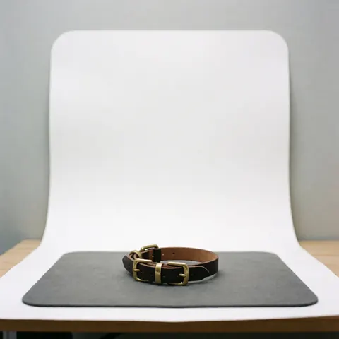 Collier en cuir marron doté de charnières en bronze, posé sur une surface grise devant un fond blanc courbe, éclairage doux et professionnel.