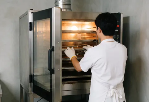 Un boulanger avec des gants ajuste un rack dans un four commercial éclairé par une lumière chaude, avec des murs en béton et de la vapeur.