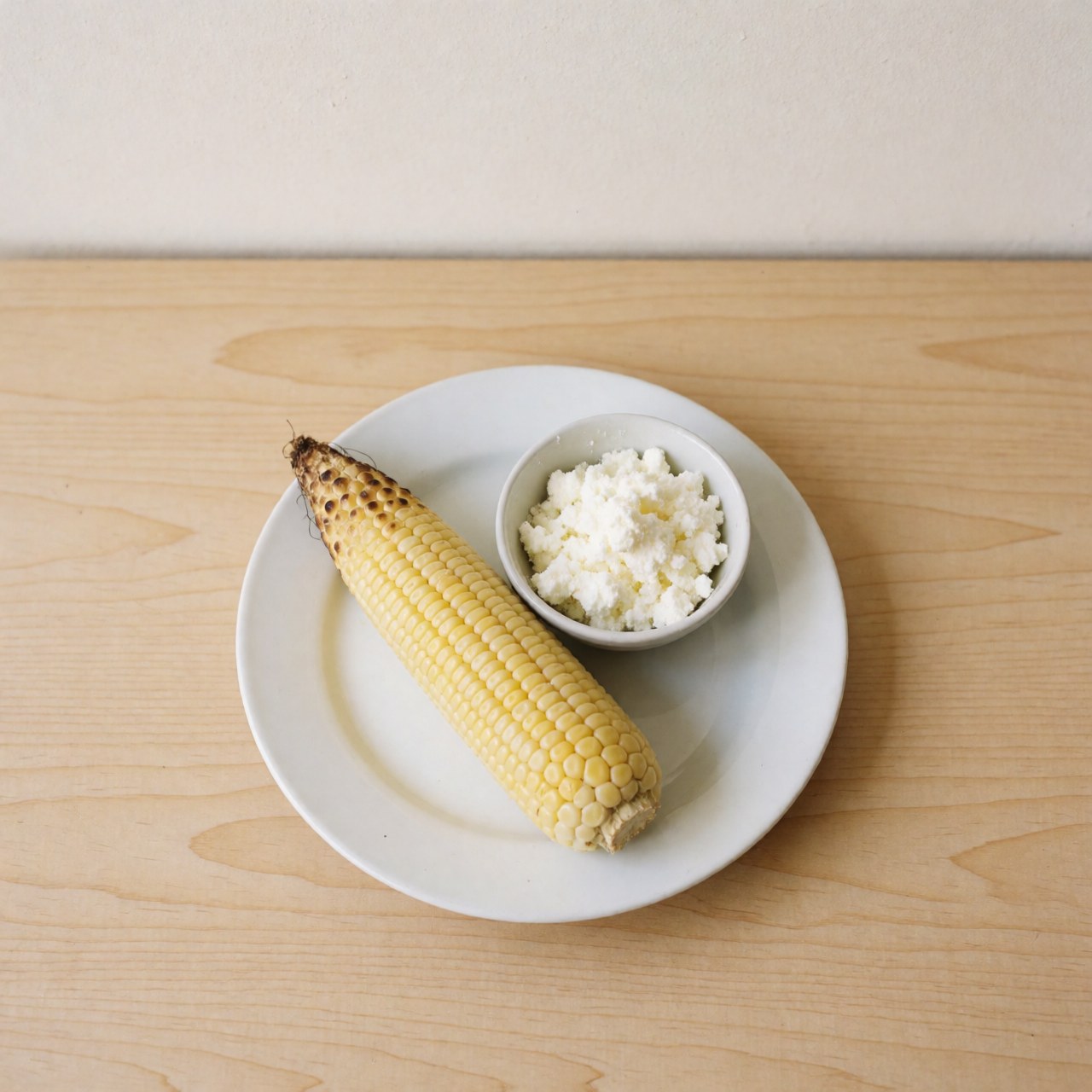 Épi de maïs péruvien cuit accompagné de fromage blanc posé sur une assiette blanche, le tout disposé sur une table en bois clair sous un éclairage naturel.
