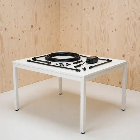 Kit d'irrigation goutte à goutte avec tuyaux noirs, émetteurs et contrôleur posés sur une table blanche devant un mur en bois clair.