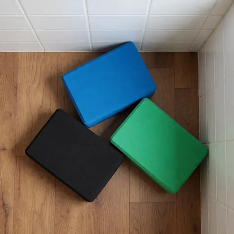 Vue de trois blocs de yoga aux couleurs bleu, vert et noir posés sur un plancher en bois clair, près d'un mur carrelé blanc.