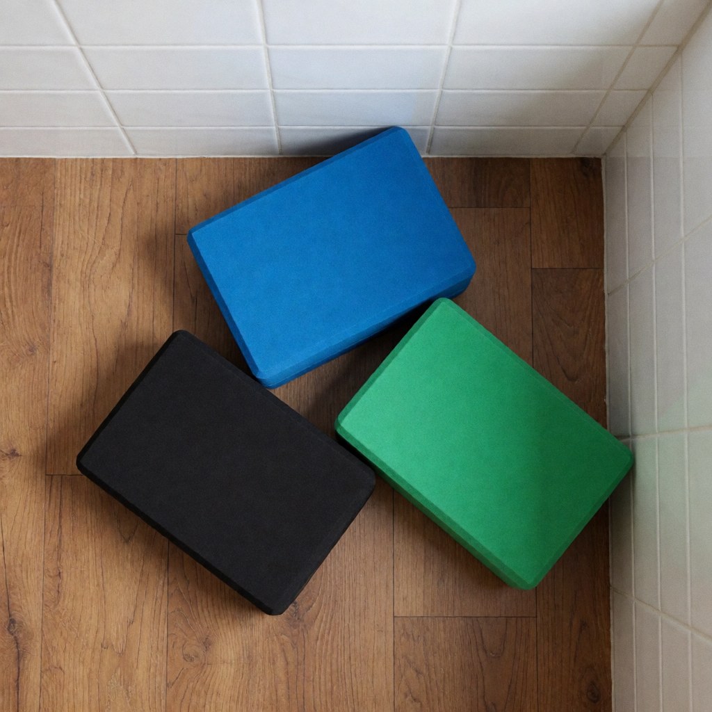 Vue de trois blocs de yoga aux couleurs bleu, vert et noir posés sur un plancher en bois clair, près d'un mur carrelé blanc.