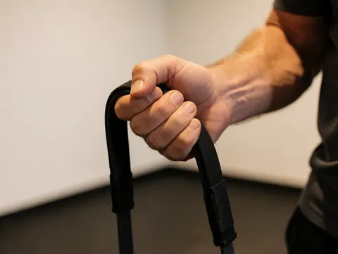 Gros plan d'une main adulte saisissant fermement une bande de résistance noire. La texture du matériau et les détails de la peau sont visibles sur un fond de mur blanc et de sol de gymnase.