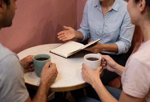 Un coach discute avec son client autour d'une petite table, tenant des mugs de café et un carnet ouvert dans une ambiance chaleureuse.