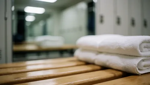 Des serviettes blanches pliées reposent sur un banc en bois dans un vestiaire de salle de sport, avec miroirs et casiers flous en arrière-plan.