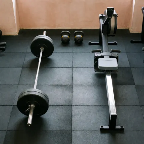 Vue de dessus d'un équipement de crossfit incluant une barre, deux kettlebells et un rameur sur sol noir avec murs terracotta, idéal pour l'entraînement.