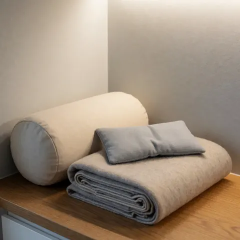 Kit de yoga restauratif composé d'un coussin, d'une couverture et d'un oreiller posés sur une table en bois, avec des murs gris et une lumière chaude.