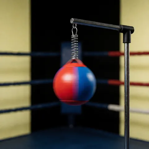 Ballon de vitesse rouge et bleu suspendu par un ressort dans un coin de ring de boxe, sur fond sombre avec des murs jaunes, pour l'entraînement des réflexes.