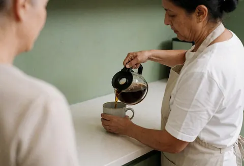 Vue arrière d'une personne versant du café d'une cafetière en verre dans une tasse sur une table blanche devant un mur vert pâle.