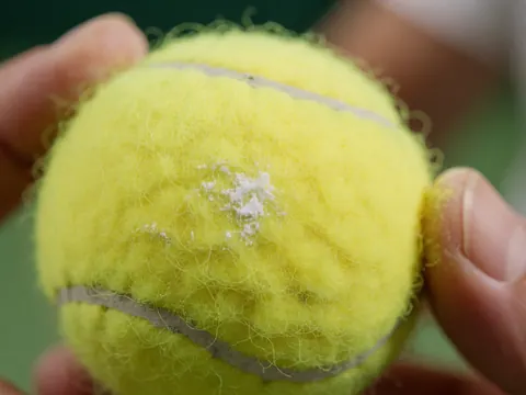 Gros plan d'une balle de tennis jaune tenue par des mains adultes, montrant la texture du feutre et la poussière blanche sur un fond vert flou.