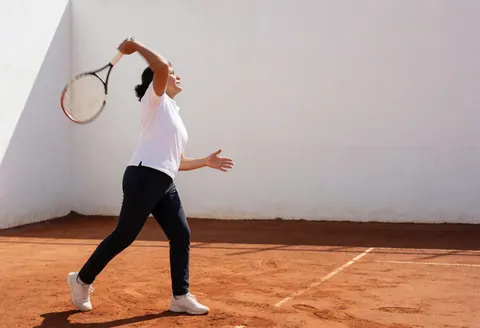 Une joueuse de tennis professionnelle en pleine action frappe un smash avec sa raquette sur un court en terre rouge devant un mur blanc.