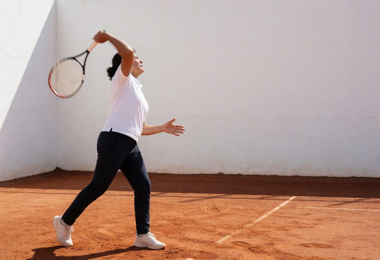 Une joueuse de tennis professionnelle en pleine action frappe un smash avec sa raquette sur un court en terre rouge devant un mur blanc.