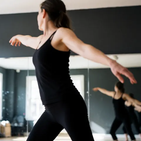 Une danseuse vêtue de noir effectue une pose expressive dans un studio aux murs gris, ses mouvements reflétés par les miroirs.