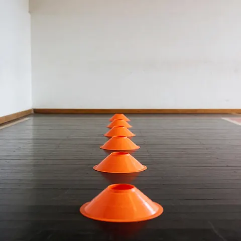 Cones orange pour l'agilité disposés en ligne sur un plancher en bois sombre avec des murs blancs en arrière-plan, idéal pour le sport.