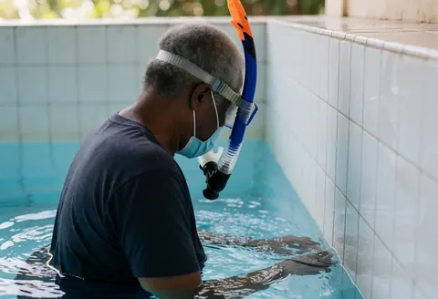 Un entraîneur de natation équipé d'un tuba et d'un masque explore le fond d'une piscine à eau claire et bleue, entouré de murs carrelés blancs sous une lumière naturelle.