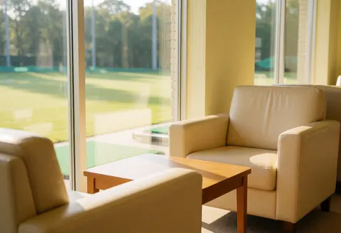 Salle d'attente de golf équipée de fauteuils beiges, d'une table en bois et offrant une vue sur le parcours. La pièce est éclairée par la lumière naturelle et dispose de murs jaune clair.