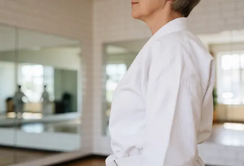 Une femme senior pratique le karate dans un dojo avec des miroirs et des murs carrelés blancs, baignée de lumière naturelle, adoptant une posture équilibrée et sereine.