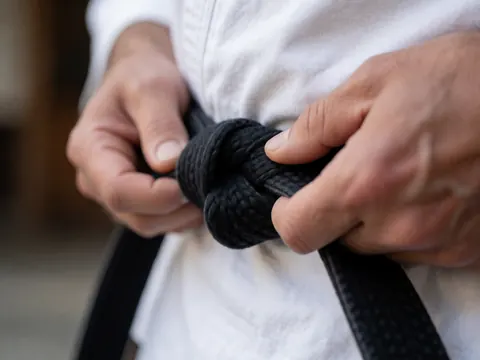 Gros plan des mains d'un adulte attachant la ceinture noire d'un uniforme de martial arts, avec texture du tissu visible et fond flou.