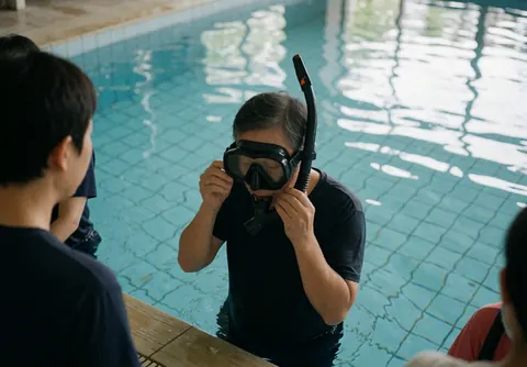 Un entraîneur de natation ajuste son masque et son tuba au bord de la piscine tandis que des étudiants observent depuis le côté.