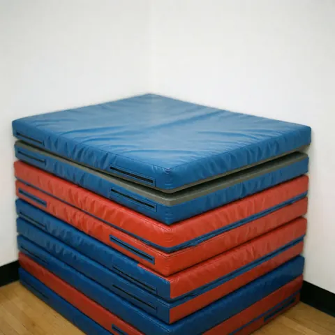 Pile de tapis rouges et bleus pour la gymnastique dans un coin de gymnase avec sol en bois et mur blanc, éclairage vif et texture visible.