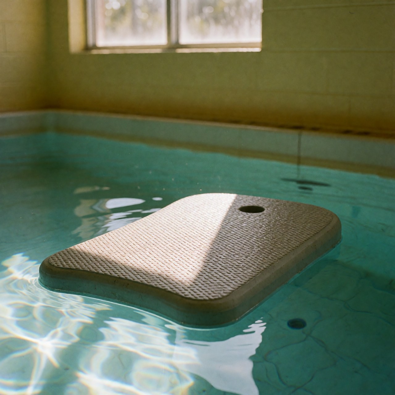 Planche de kickboard antidérapante flottant sur l'eau claire d'une piscine intérieure, éclairée par la lumière du soleil créant des reflets, avec des murs jaunes en arrière-plan.