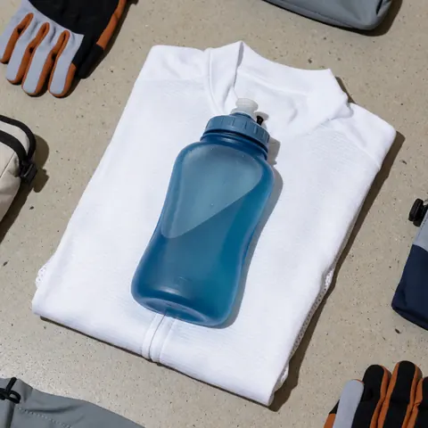 Une gourde bleue posée sur un maillot de cyclisme blanc plié, accompagnée de gants et d'un sac sur un sol en béton pour l'entraînement.