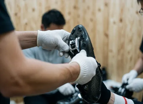 Des mains gantées ajustent une cale en carbone sur une chaussure de cyclisme, avec des murs en bois en arrière-plan et d'autres personnes travaillant au loin.