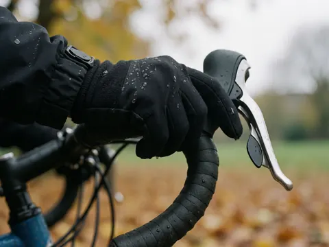 Gros plan d'une main portant un gant noir agrippant fermement le guidon d'un vélo, avec des gouttes de pluie visibles sur les feuilles mortes en arrière-plan flou.