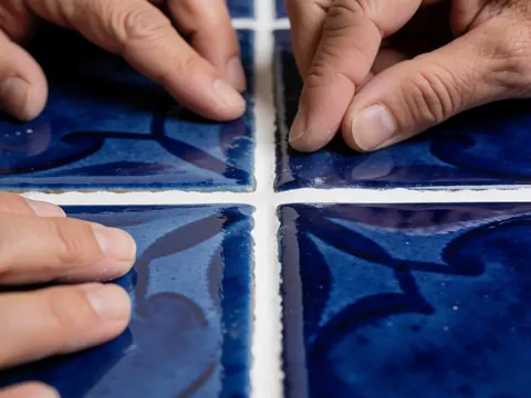Mains d'adulte ajustant du mortier blanc entre des carreaux de céramique bleus à motifs, avec une lumière douce mettant en valeur les détails brillants.