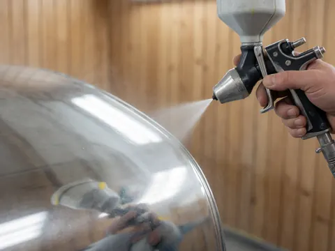 Une main tient un pistolet à peinture pour appliquer une finition brillante sur une surface métallique courbe dans un atelier aux murs en bois.
