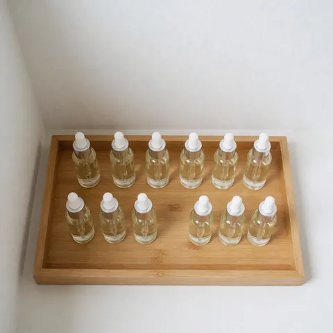 Dix bouteilles en verre avec compte-gouttes disposées en deux rangées sur un plateau en bois clair, fond blanc minimaliste pour soins corporels.