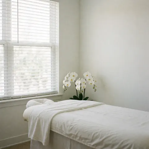 Intérieur minimaliste d'un spa avec une table de massage blanche, des orchidées blanches et la lumière naturelle filtrant par les stores.