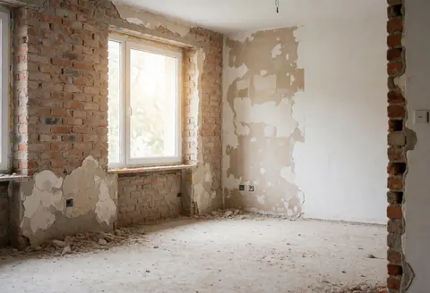 Salle en cours de rénovation présentant des murs en briques exposées, un sol poussiéreux et des fenêtres laissant entrer la lumière naturelle.