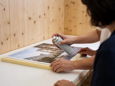 Une main applique un adhésif en spray sur une impression graphique fixée sur un panneau en mousse dans un atelier.