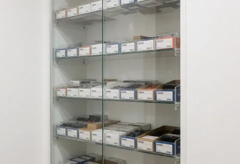 Vitrine en verre contenant des étagères remplies de boîtes de pièces électroniques sur fond blanc avec un éclairage uniforme pour une boutique de réparation.