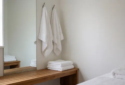 Cabine privée et propre équipée d'un banc en bois, d'un grand miroir et de serviettes blanches pliées, baignée d'une lumière naturelle douce.