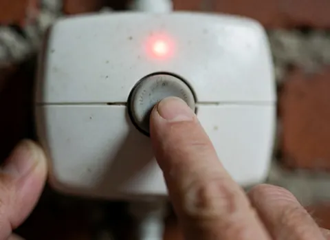 Gros plan d'un doigt en train d'appuyer sur un bouton de contrôle émettant une lumière rouge, avec un mur de briques en arrière-plan pour le détail.
