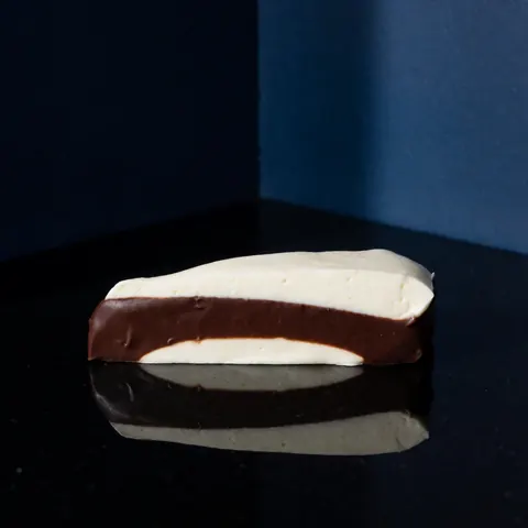 Gâteau au chocolat avec crème sur une surface noire réfléchissante, éclairage dramatique, fond bleu foncé, textures douces et couches visibles.