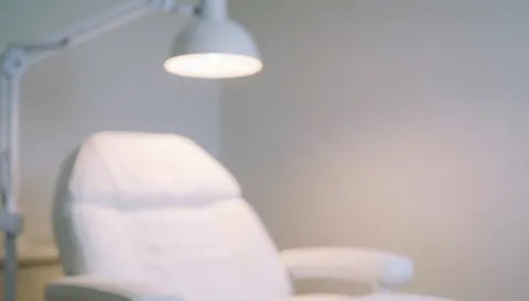 Arrière-plan flou d'une salle de traitement avec chaise blanche et lampe murale, ambiance sereine et minimaliste pour le microblading, couleurs pastel et lumière chaude.