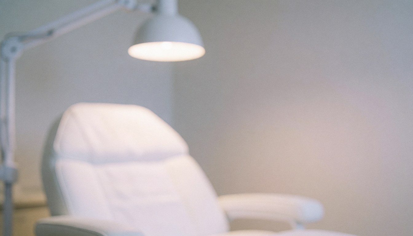 Arrière-plan flou d'une salle de traitement avec chaise blanche et lampe murale, ambiance sereine et minimaliste pour le microblading, couleurs pastel et lumière chaude.