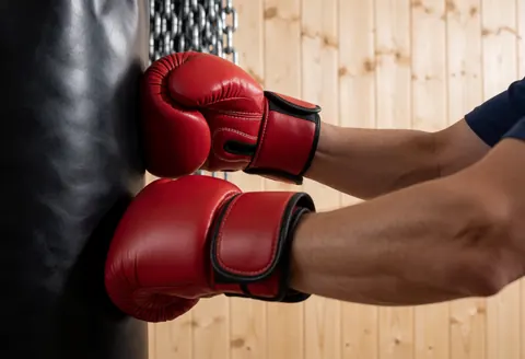 Gros plan sur des mains portant des gants rouges qui impactent un sac de boxe noir, avec des chaînes et un mur en bois en arrière-plan sous une lumière dramatique.